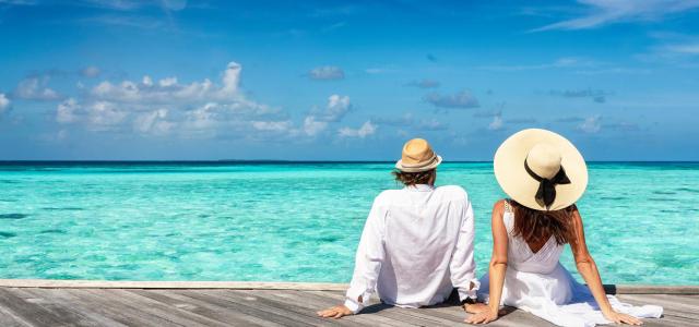 A beautiful couple in white summer clothing sits on a wooden pier and enjoys the view to the turquoise ocean of the tropical Maldives islands